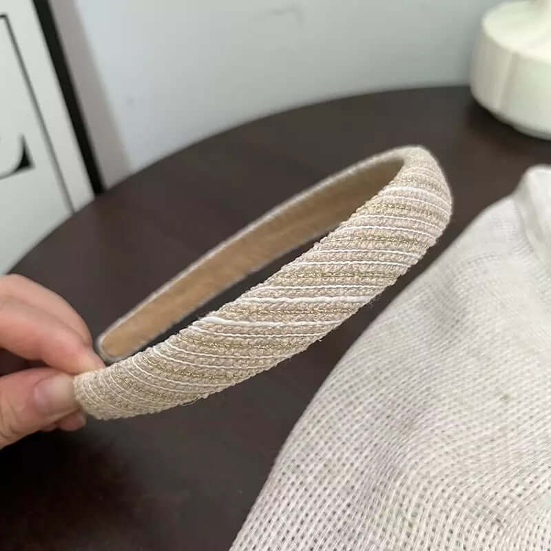 A single cream and gold metallic woven textured headband held by hand on a dark wood table.