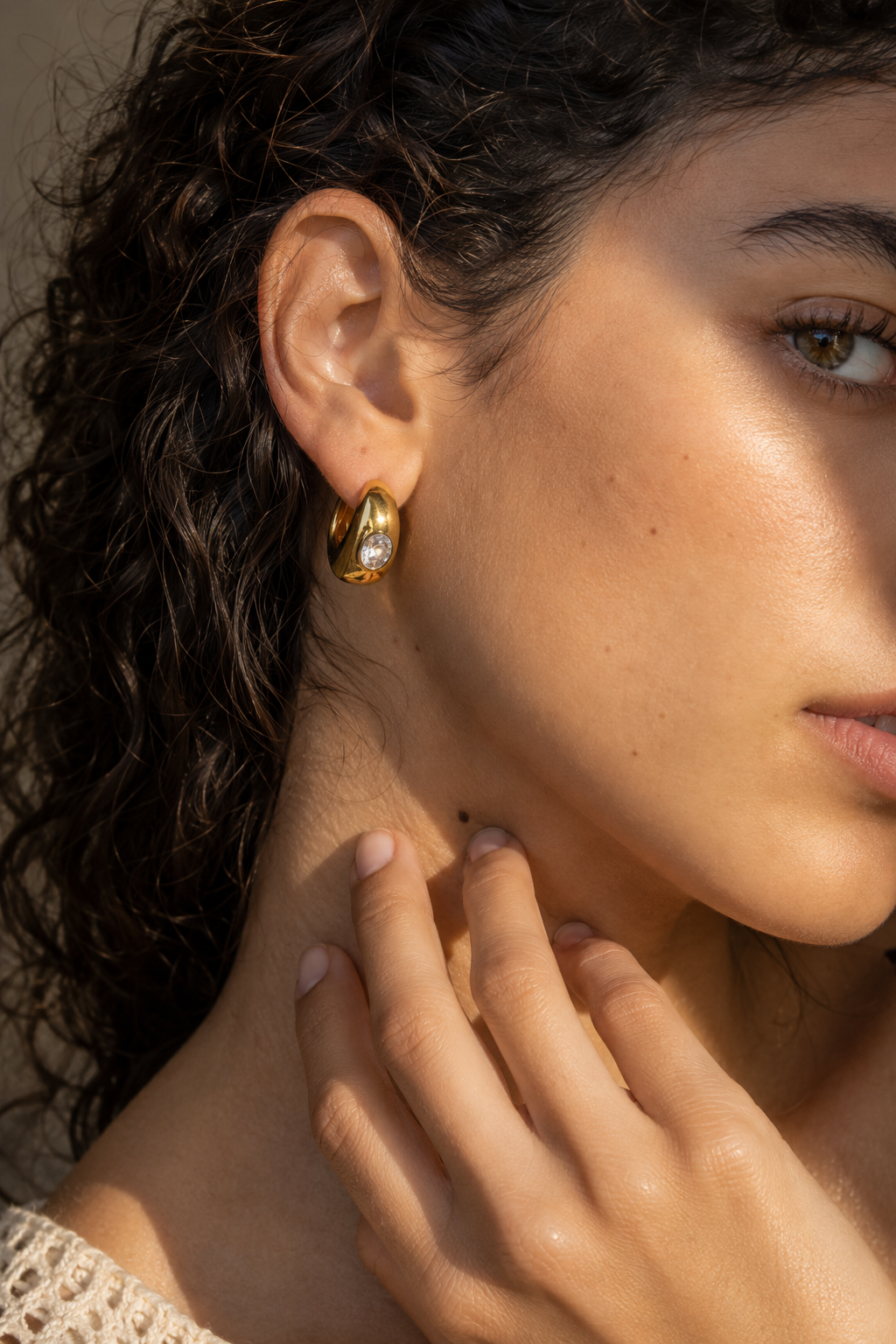 Close-up of a model with curly hair wearing the gold Cascade EARRINGS, a chunky huggie hoop with a bezel-set white stone.
