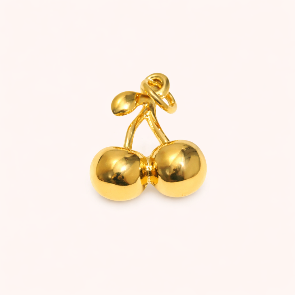The Cherry CHARM in a studio flat lay, a polished gold pendant shaped like a pair of cherries on a cream background.