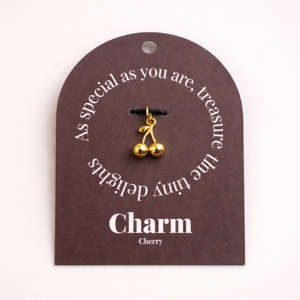 The Cherry CHARM displayed on a brown Charm gift tag, the gold cherry pendant hanging from a black cord.