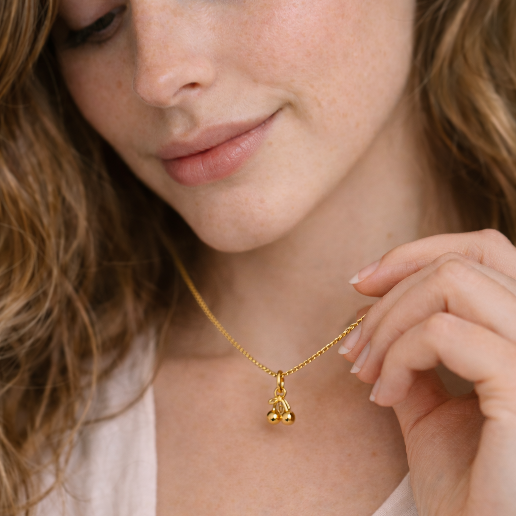 The Cherry CHARM worn at the neckline, a polished gold cherry pendant on a fine gold chain held between two fingers.