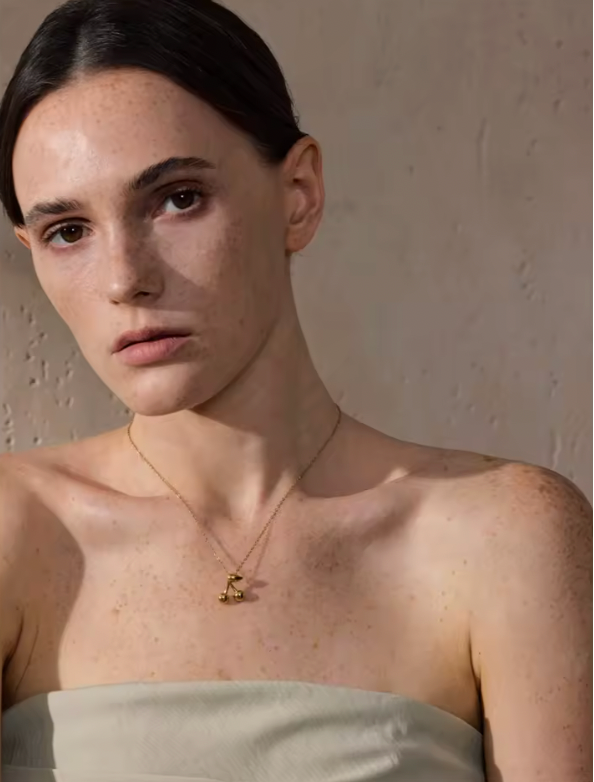 The Cherry On Top NECKLACE worn at the neckline, a small gold-toned cherry pendant on a fine chain against warm-lit skin.