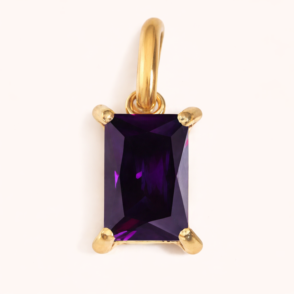Close-up of the Classic Drop Charm in a deep purple amethyst stone, set in a gold prong mount on a cream background.