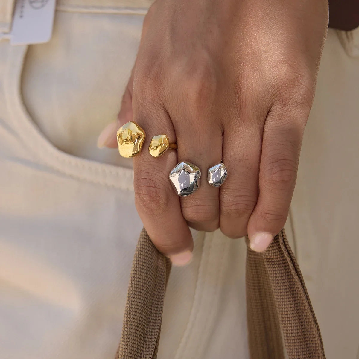 Hand holding a woven bag strap, wearing gold and silver-tone Cloud RINGS against cream trousers — Folie à Trois
