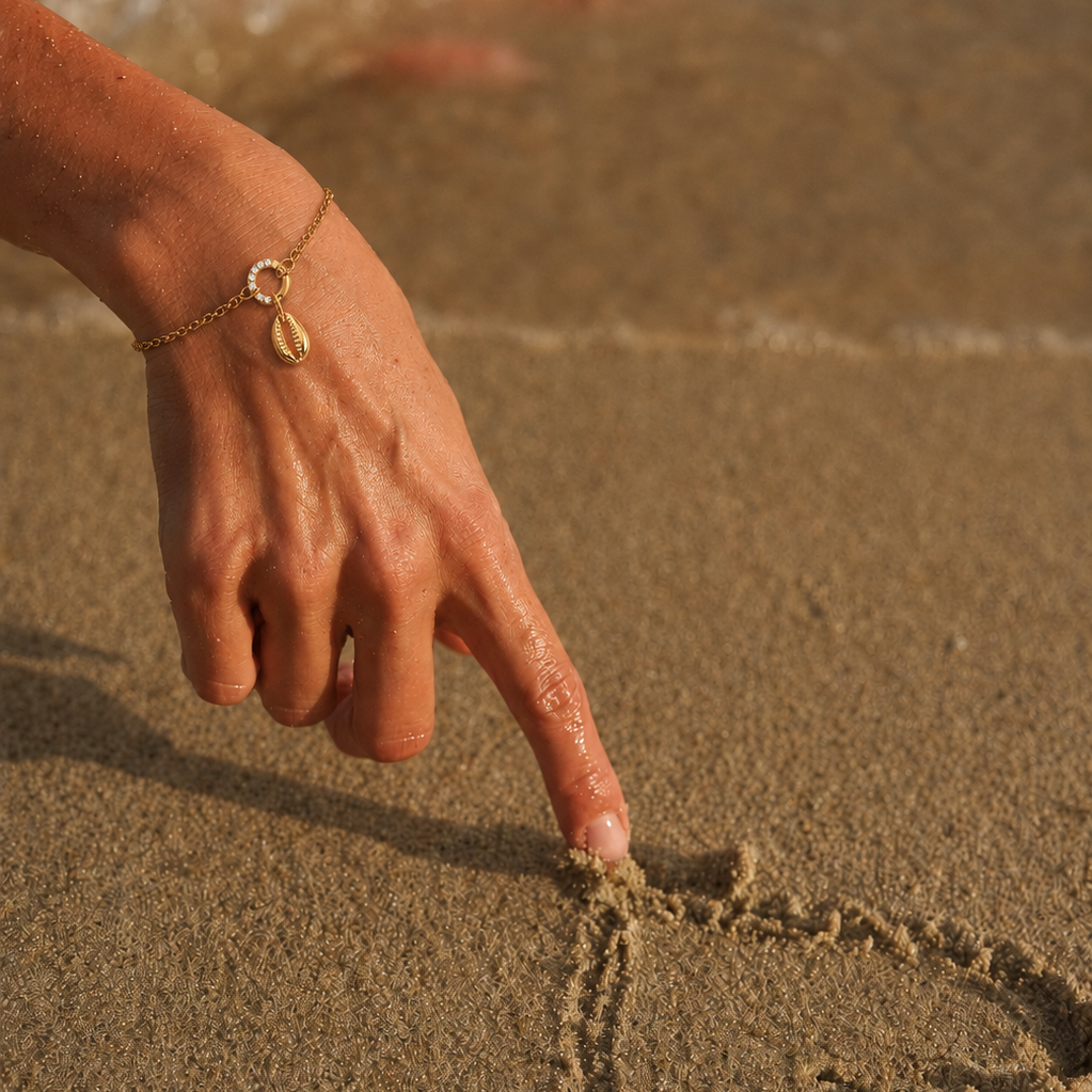 The Conch Shell CHARM worn on a gold bracelet with a pearl and crystal clasp, resting on a hand tracing wet sand at the beach.