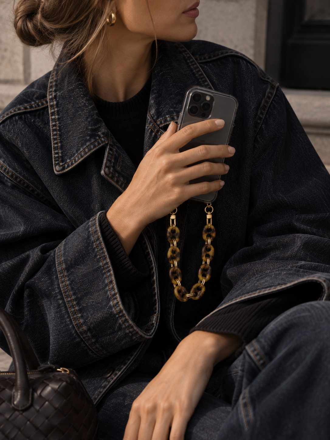 Folie à Trois Corvin SHORT PHONE CHAIN in tortoiseshell coin links, clipped to a phone held against a denim jacket with a woven bag nearby.