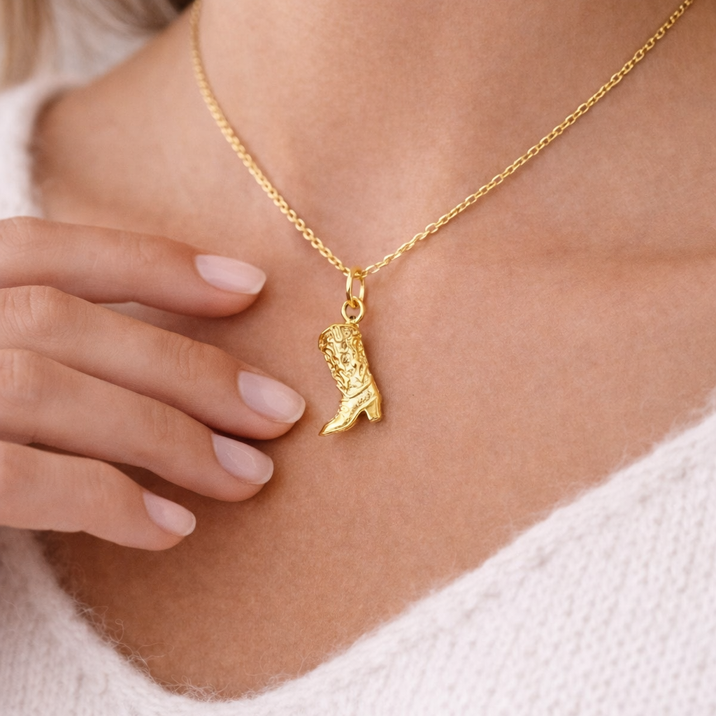 The Cowboy CHARM worn at the neckline, a textured gold cowboy boot pendant on a fine chain against a cream sweater.