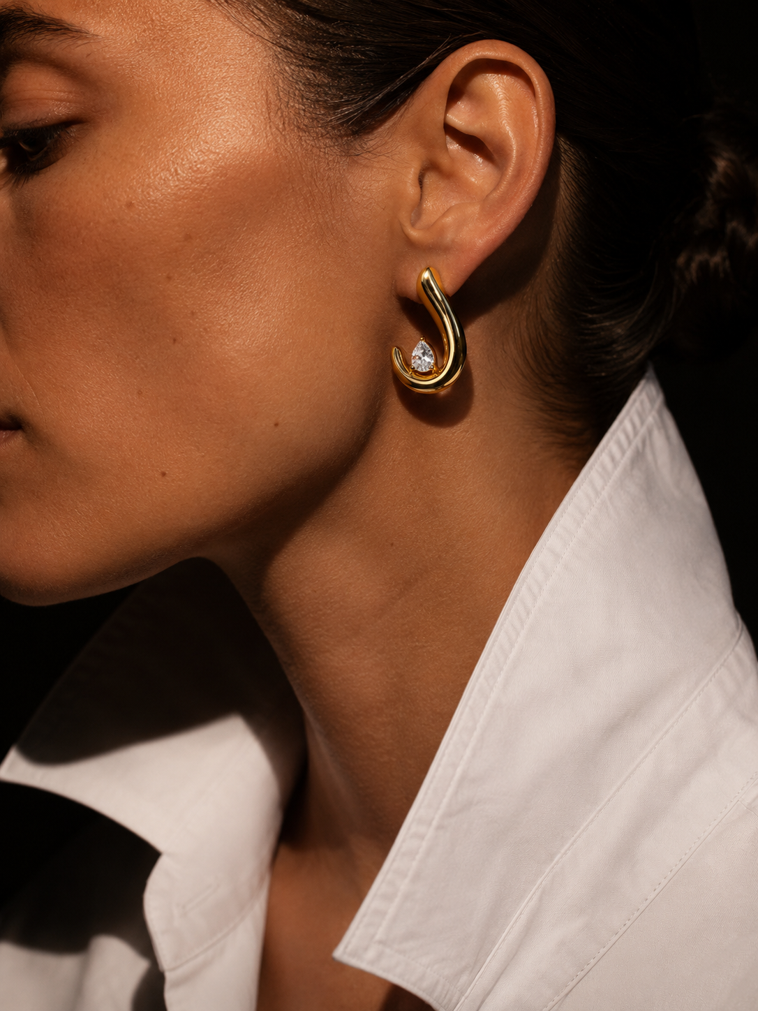 Close-up of a model in a white shirt wearing the gold Crescent EARRINGS, a curved hoop set with a pear-cut zirconia.
