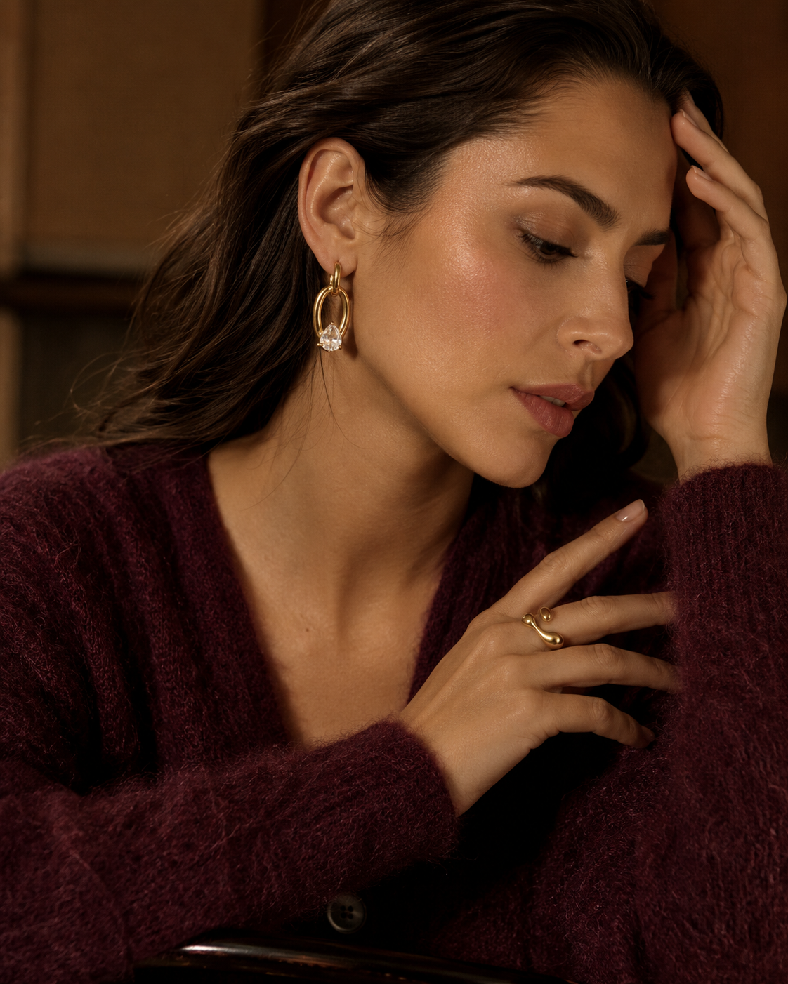Model in a burgundy sweater wears the gold Curve EARRINGS, an oval chain-link drop set with a pear-cut white stone.