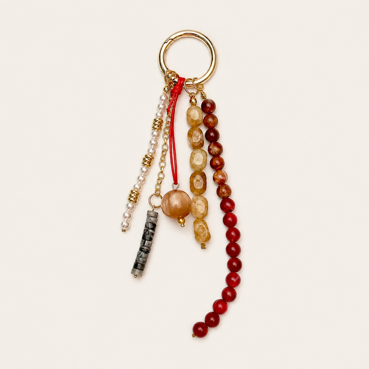 Folie à Trois Delphine BAG CHARM, a beaded tassel with pearl, grey, tan, and deep red bead strands on a gold hoop, shown on a cream background.