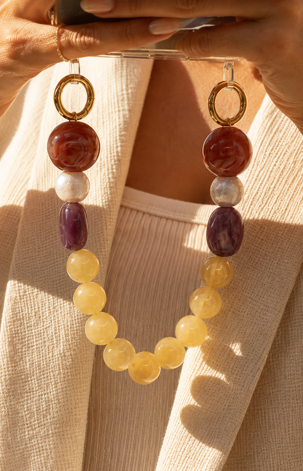 Sunlit close-up of a woman in a cream blazer holding her phone by a red, purple, and yellow bead chain — Folie à Trois Delphine phone chain