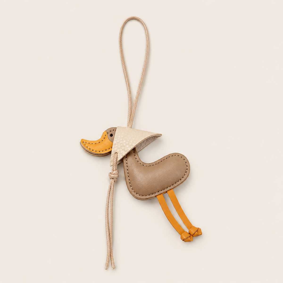 Taupe leather duck-shaped bag charm with a cream cord loop and yellow beak and feet, Folie à Trois Duckling BAG CHARM on a neutral background.