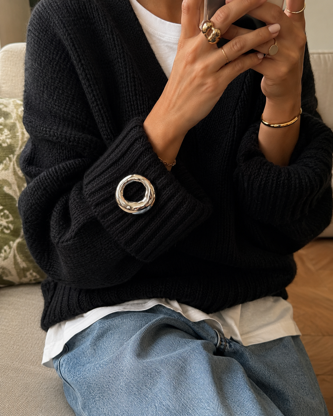Duna BROOCH in silver, a molten organic ring shape cinching the sleeve cuff of a black knit sweater, worn while holding a phone.