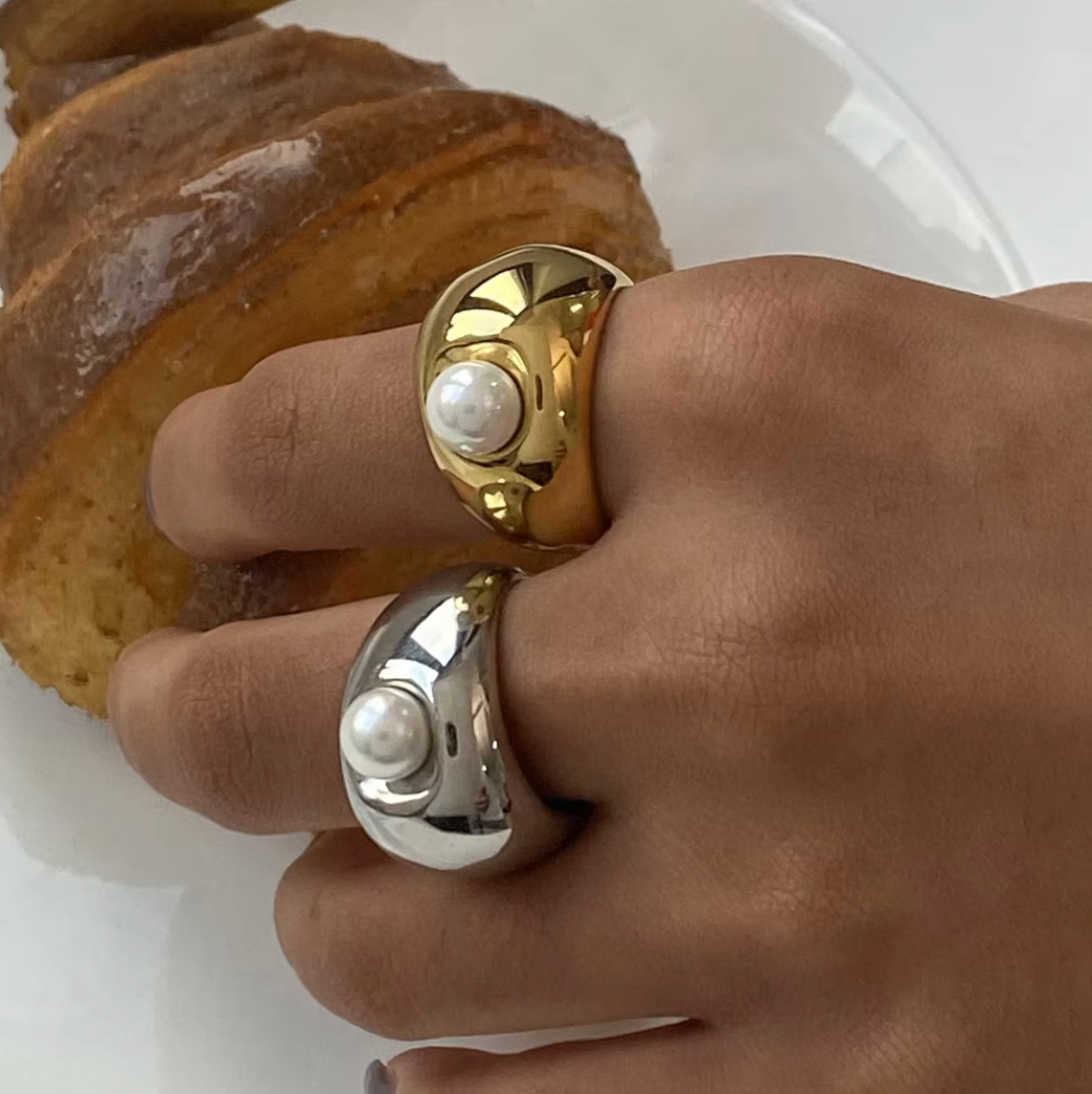 Close-up of a hand stacked with the gold and silver chunky dome Elka RING styles, each set with a pearl, near a croissant.