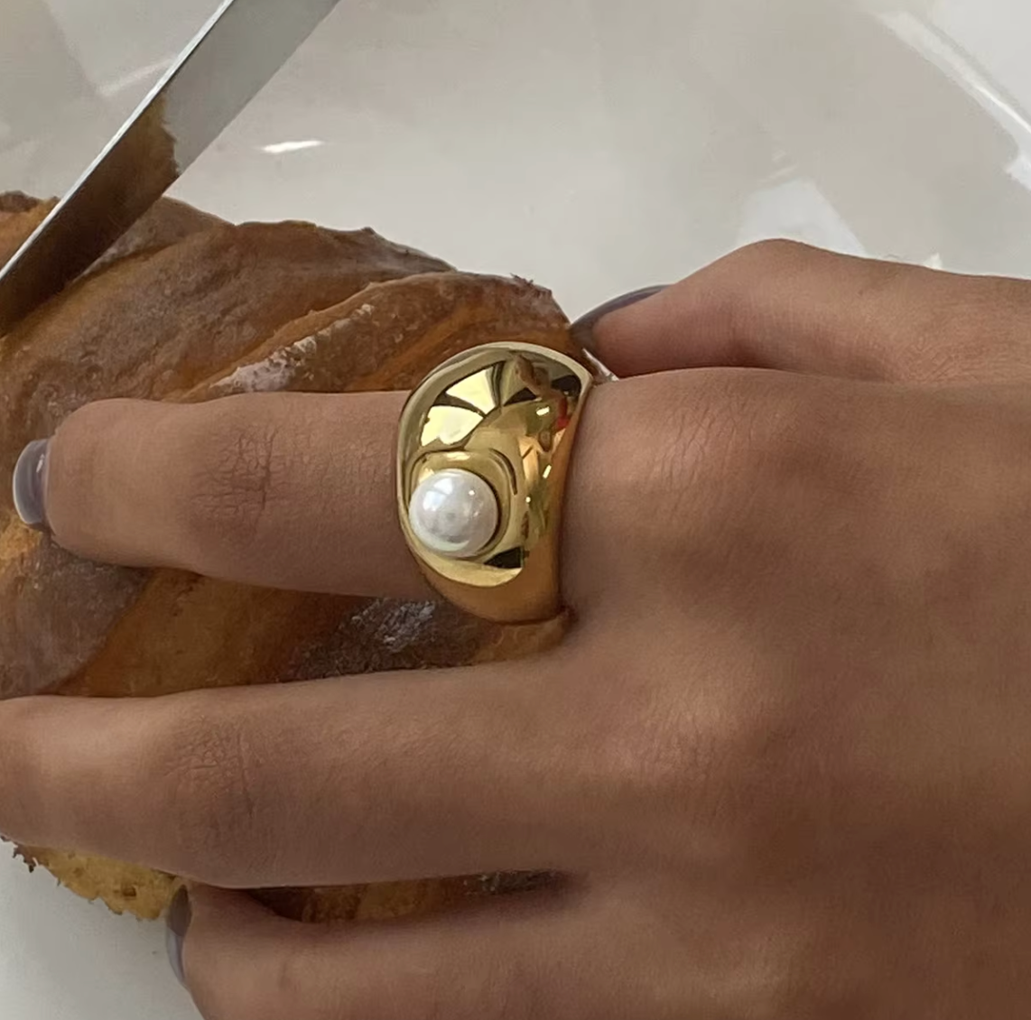 Close-up of a hand slicing bread while wearing the chunky gold dome Elka RING set with a single pearl.