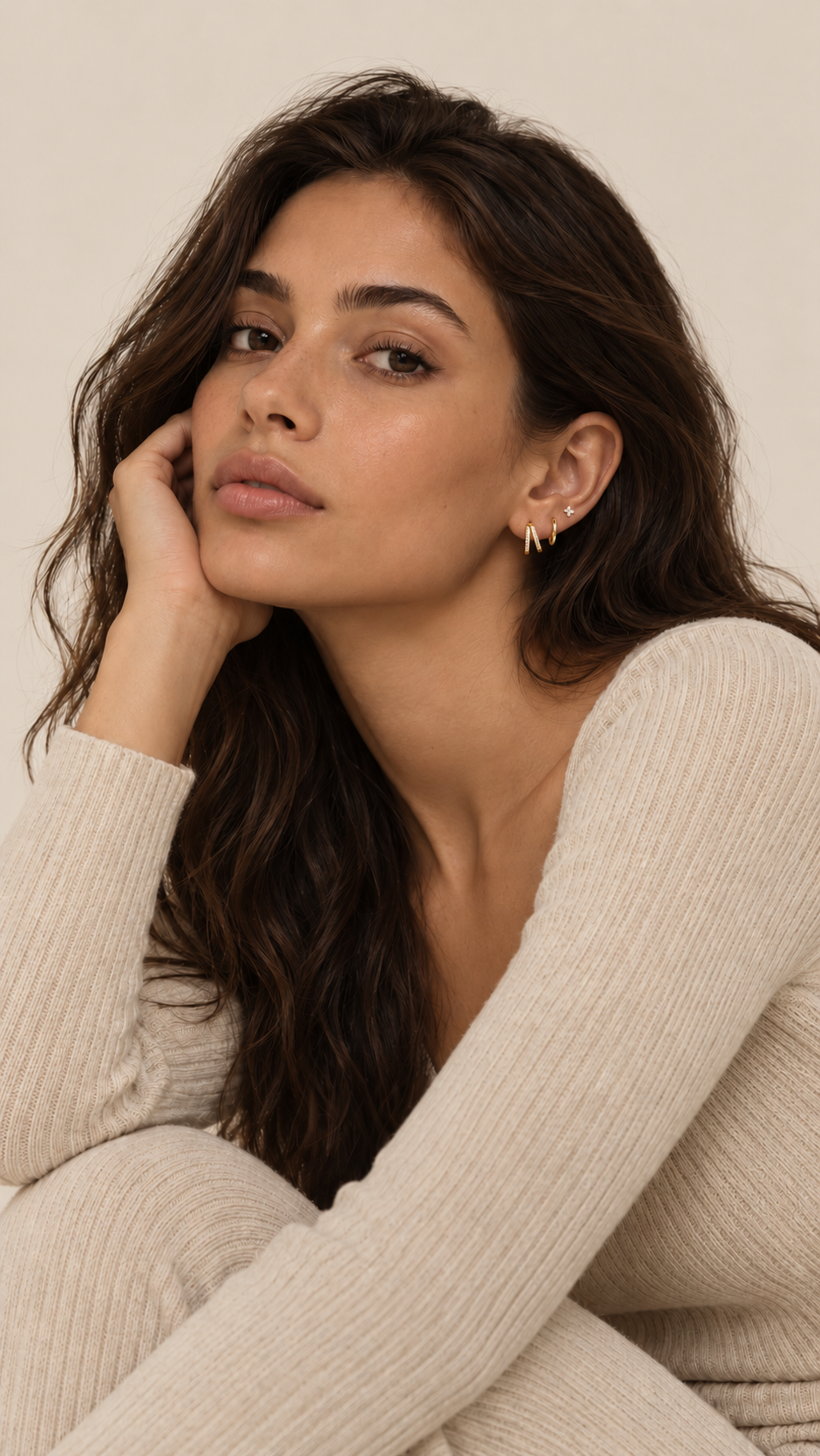 Dark-haired model resting her chin on her hand, wearing the gold Faye SET hoops and a four-point crystal stud, in a cream knit.