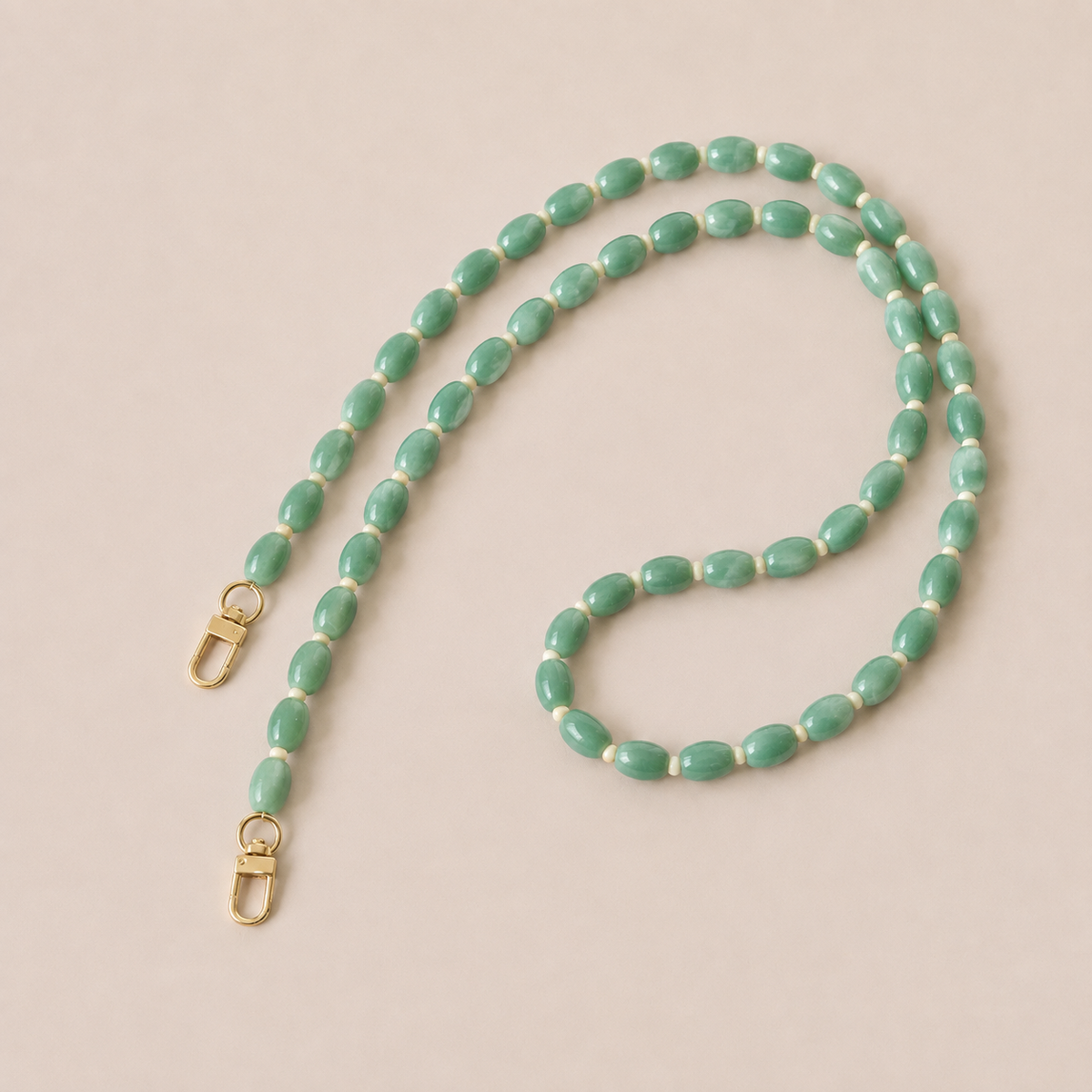 Folie à Trois Fennel long phone chain in jade green oval beads with cream spacers and gold clasps, laid flat on a cream background.