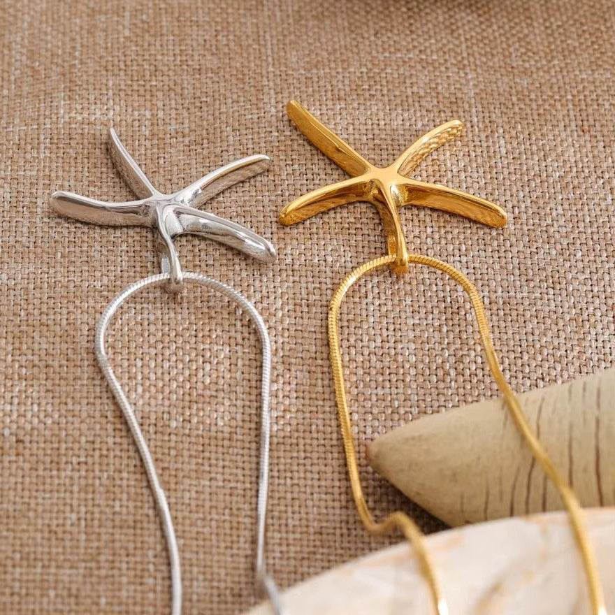 The Fiji NECKLACE shown in gold and silver side by side, both starfish pendants draped over a textured shell on woven fabric.