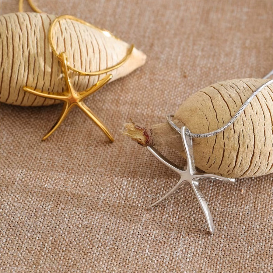 The Fiji NECKLACE shown in gold and silver from a different angle, both starfish charms resting against woven fabric and shells.