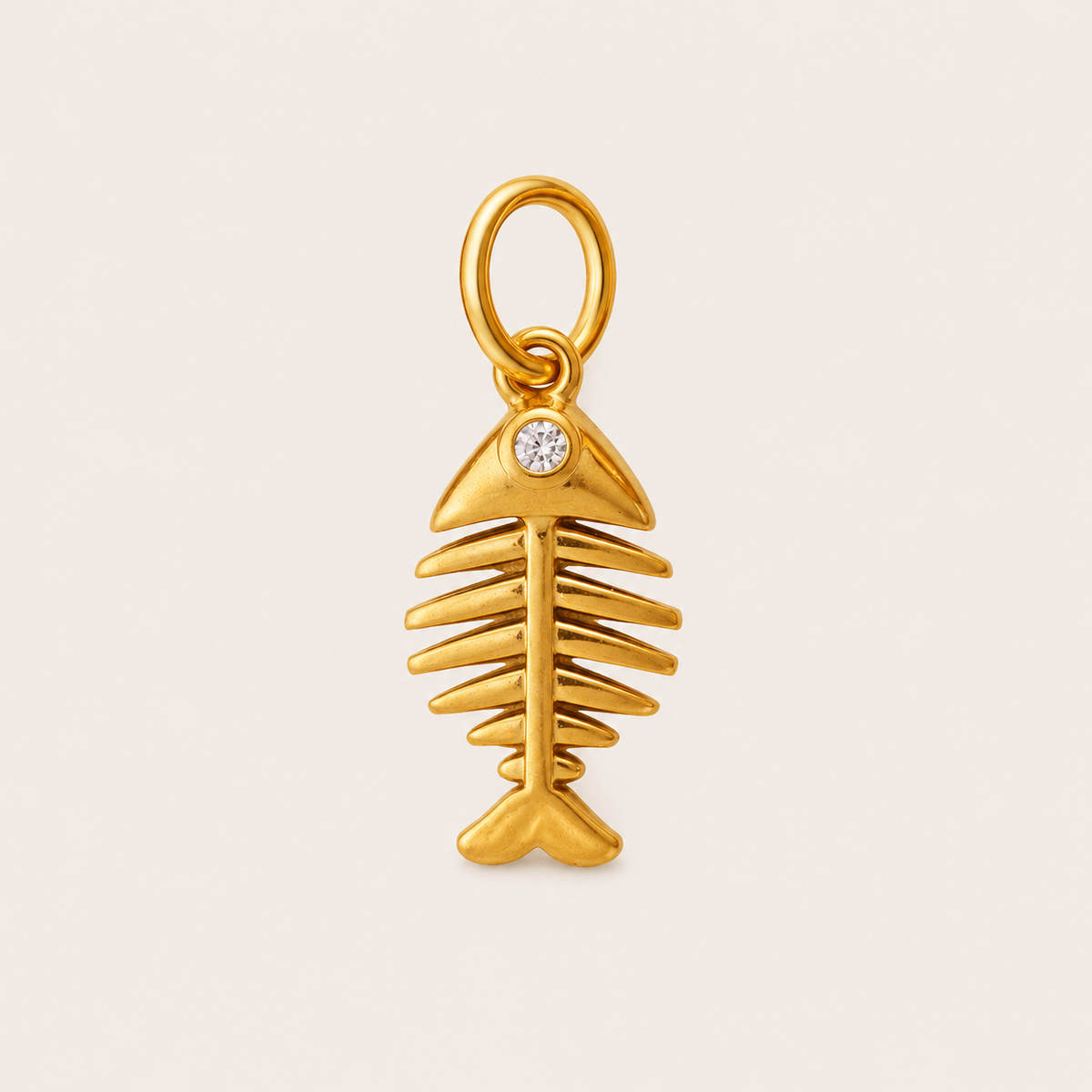 The Fishbone CHARM in gold, a fish skeleton pendant with a small clear rhinestone eye, shown on a light beige background.