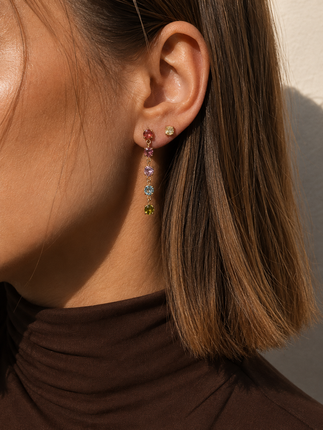 Close-up of a model in a brown turtleneck wearing the gold Fringe EARRINGS, graduated pink, ruby, yellow and green stones.