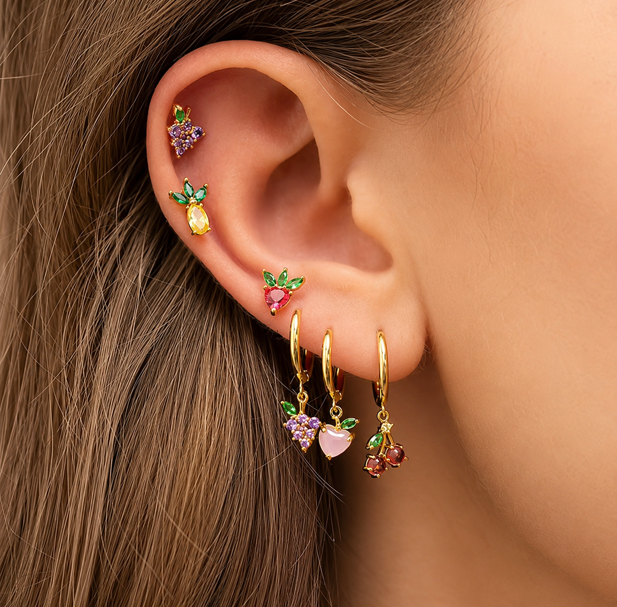 Close-up of a model's ear stacked with five tiny gold fruit-charm studs from the Little Fruit EARRINGS in yellow, pink, and purple stones.