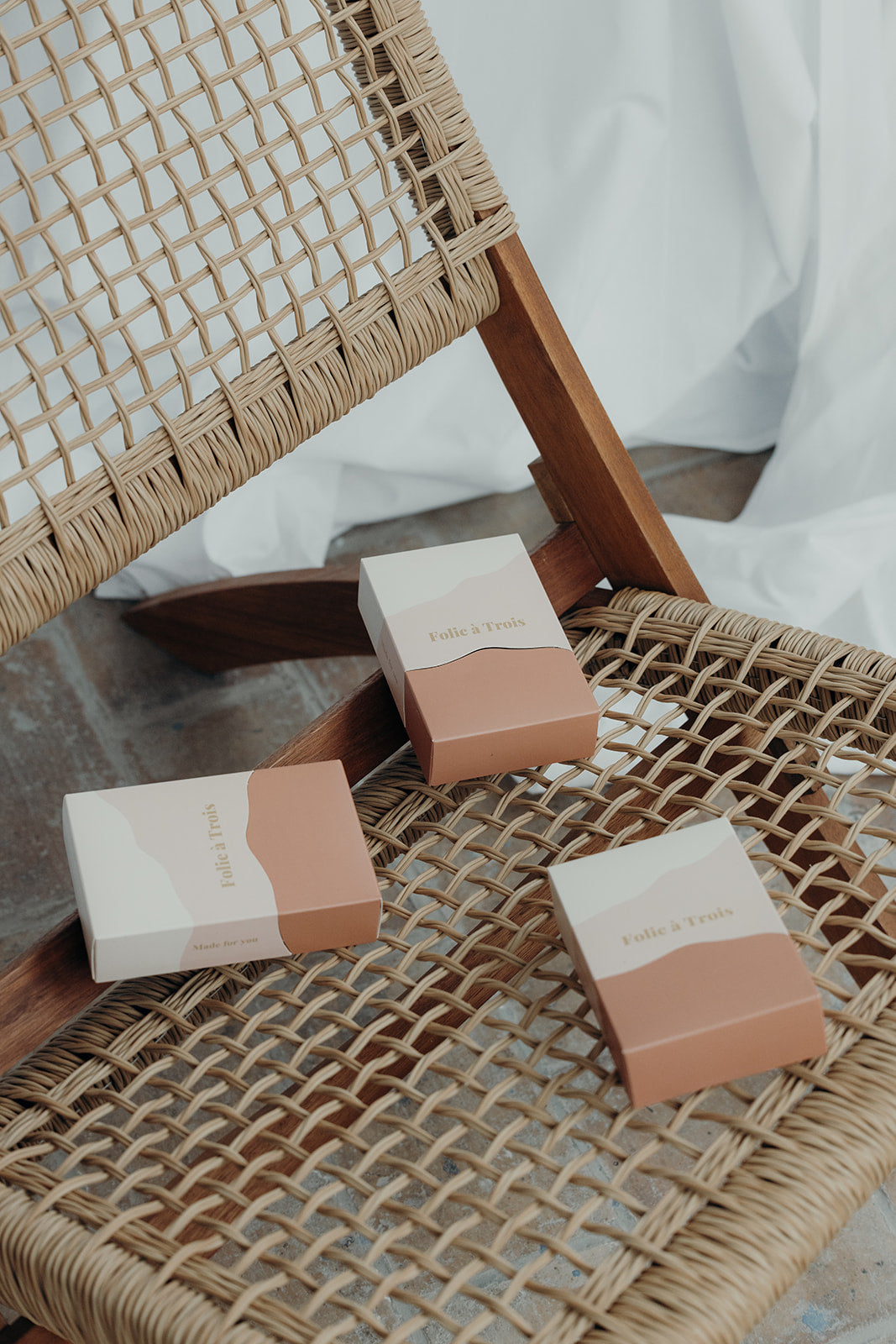 Three Folie à Trois Gift Box packages in cream and terracotta scattered across the seat of a woven rattan lounge chair.