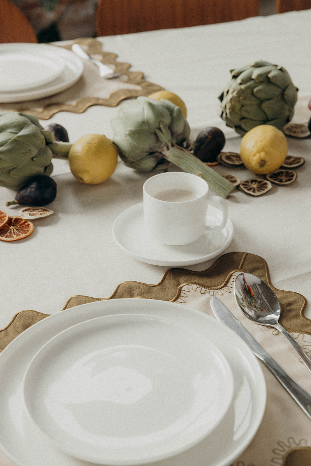 Table setting with white plates, cutlery, and a coffee cup arranged on the olive Gisèle Scallop placemat with a fruit garland nearby.