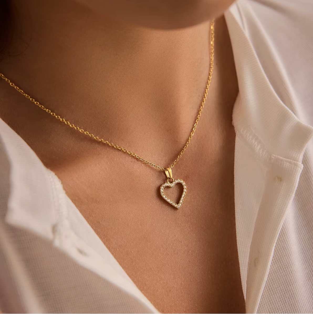 The Golden Heart NECKLACE worn at the neckline, a pave crystal open-heart pendant on a gold chain against a white shirt.