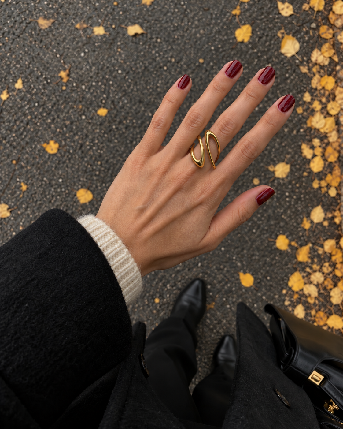 Hand with burgundy nails over autumn leaves wears the gold Infini RING, an open figure-eight spiral — Folie à Trois wholesale