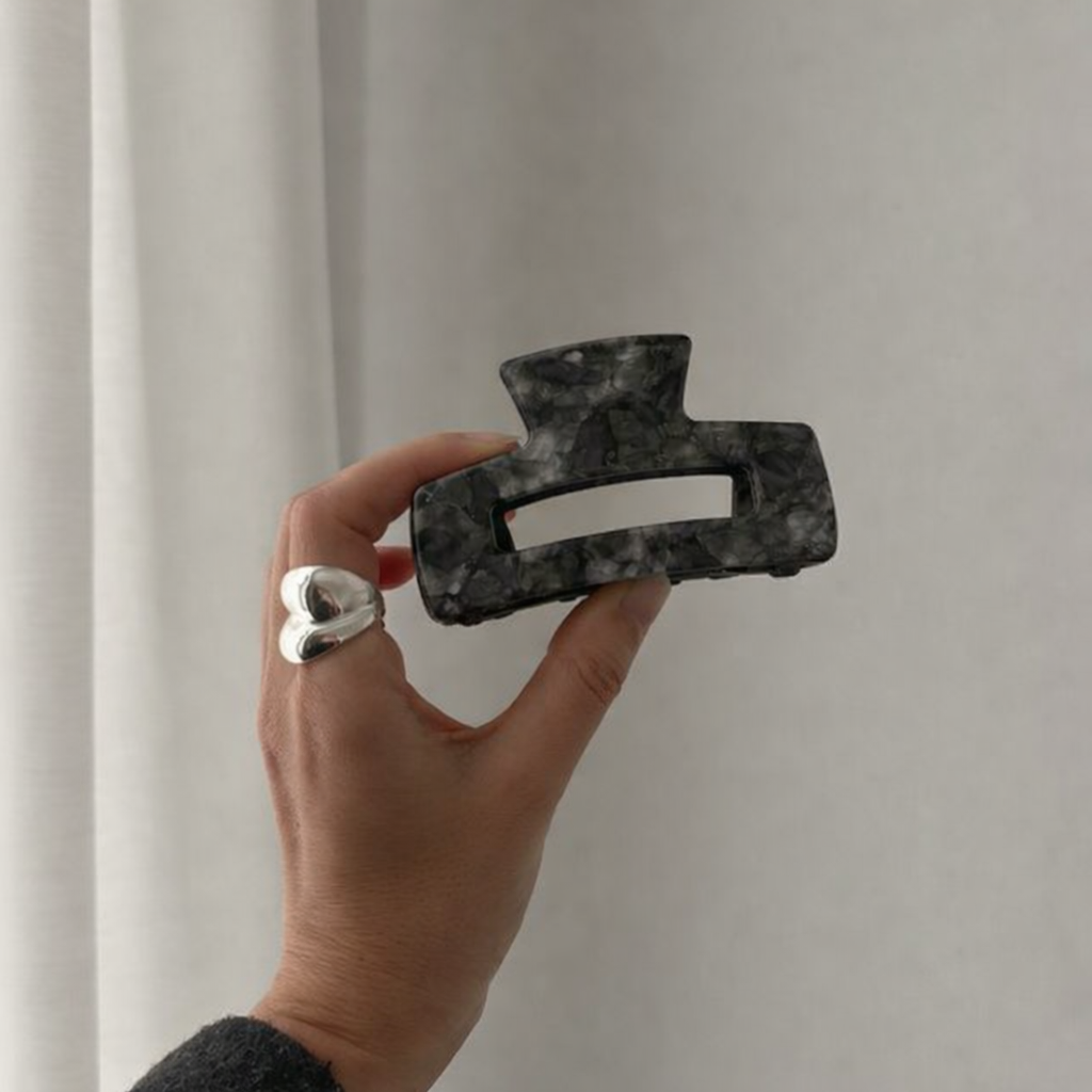 It Girl Big HAIR CLIP's charcoal grey marbled claw clip held up by hand against a curtain, styled with a silver heart ring.