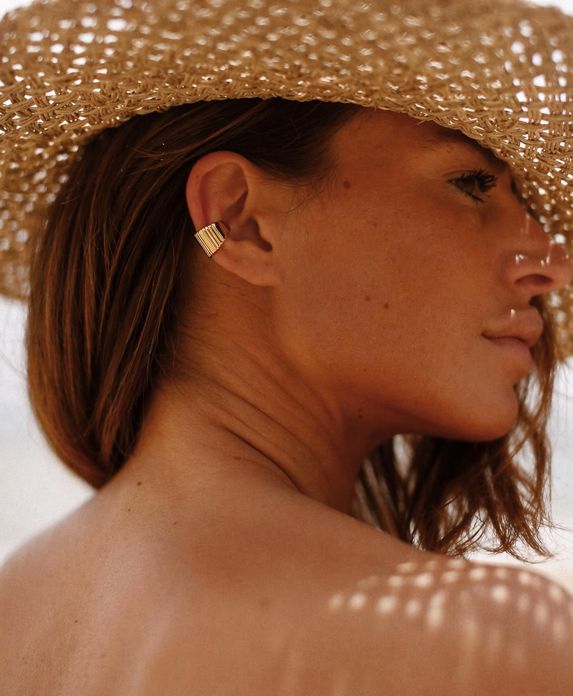 A model wears a gold Ivette EAR CUFF with a fluted, ribbed texture on her ear, styled with a woven straw hat.