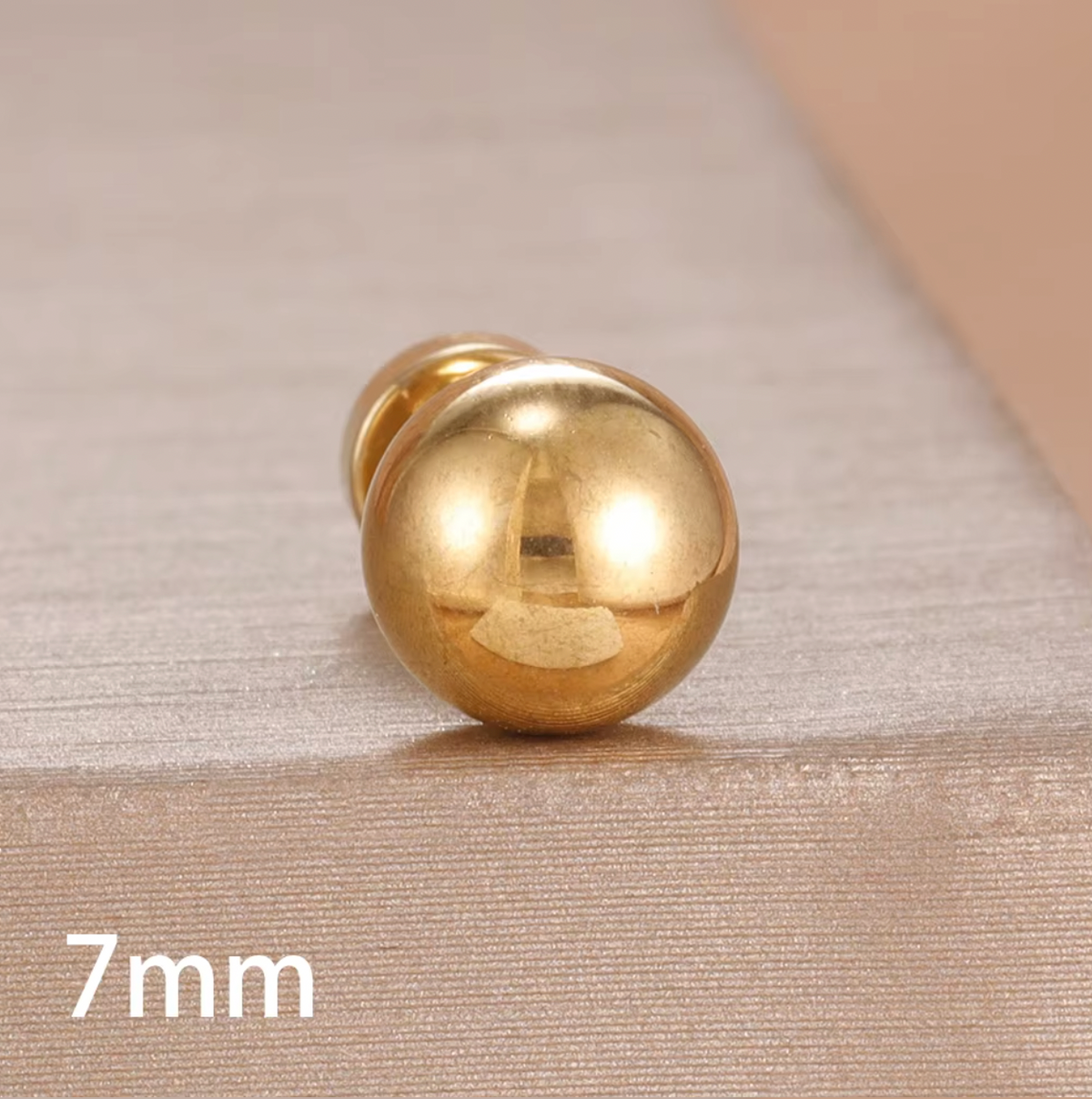 The Jean PIERCING in gold, a single 7mm labret stud shown in close-up on a textured tan surface.
