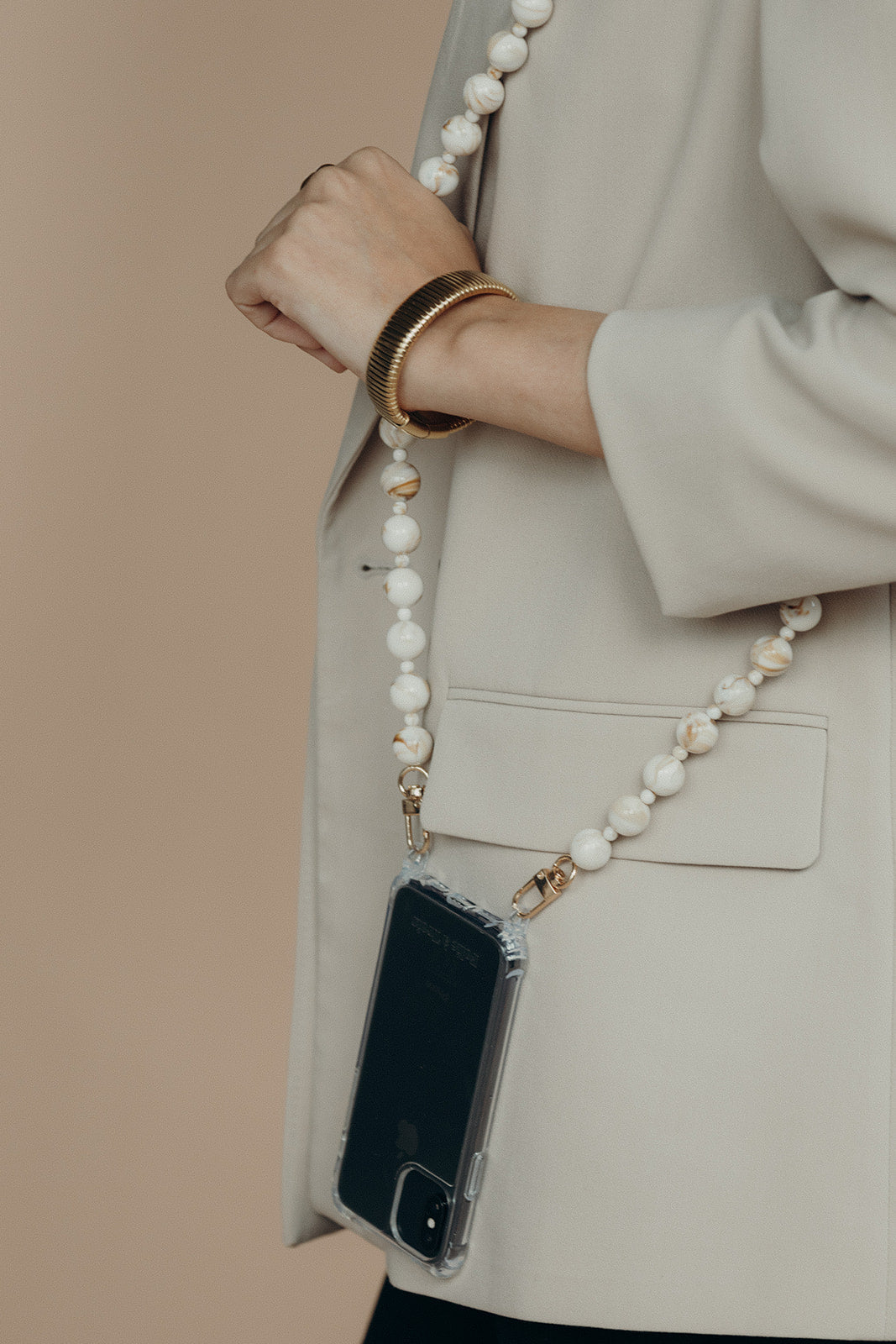 A woman holding a phone by its cream and gold marbled bead phone chain, worn crossbody over a beige blazer.