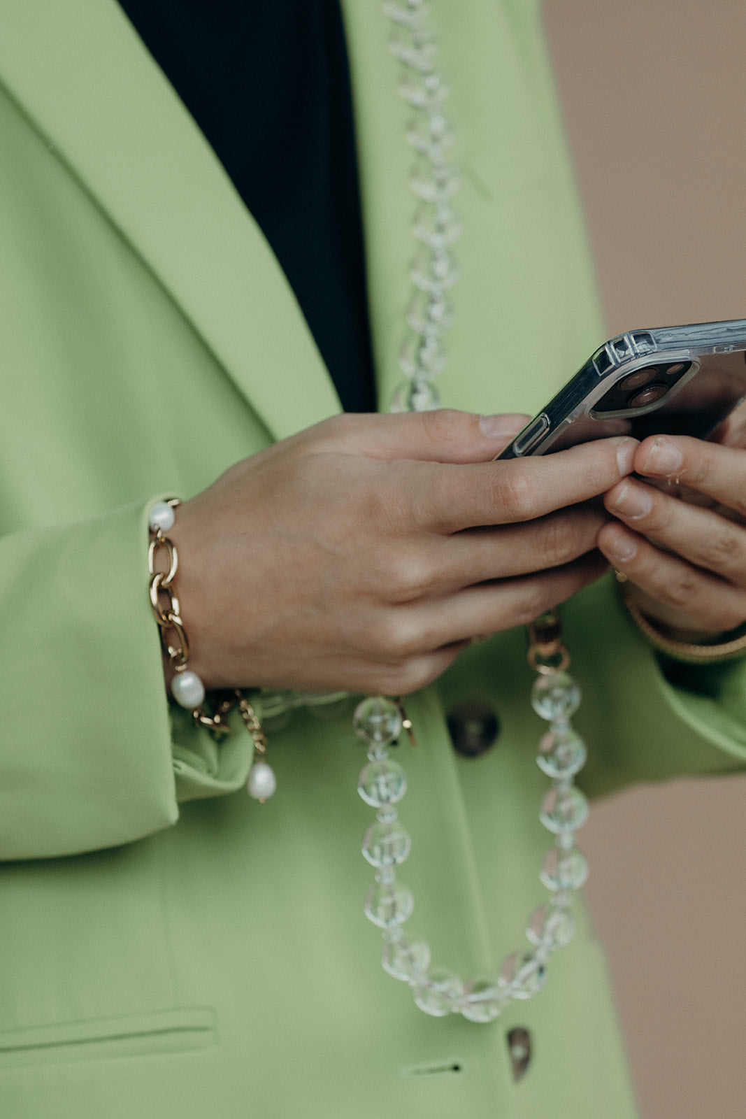 Close-up of a woman on the phone, wearing a clear glass bead phone chain and a gold chain bracelet with pearl charms, over a green blazer.