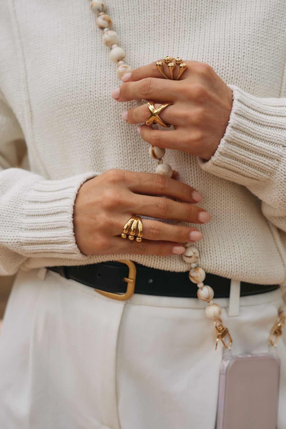 Close-up of hands wearing statement gold rings, resting on a cream and gold marbled bead phone chain over a cream sweater.