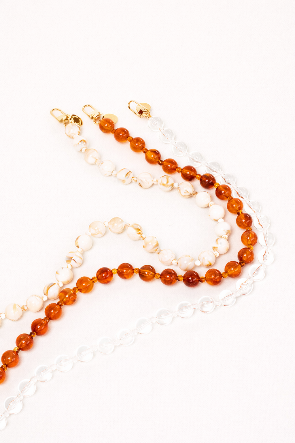 Three bead phone chains in cream and gold marble, amber, and clear glass, laid out together with gold clasps.