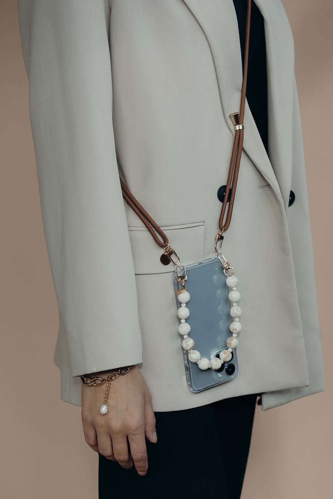 A phone in a blue case with a white and black beaded strap and a brown cord strap, worn against a beige coat.