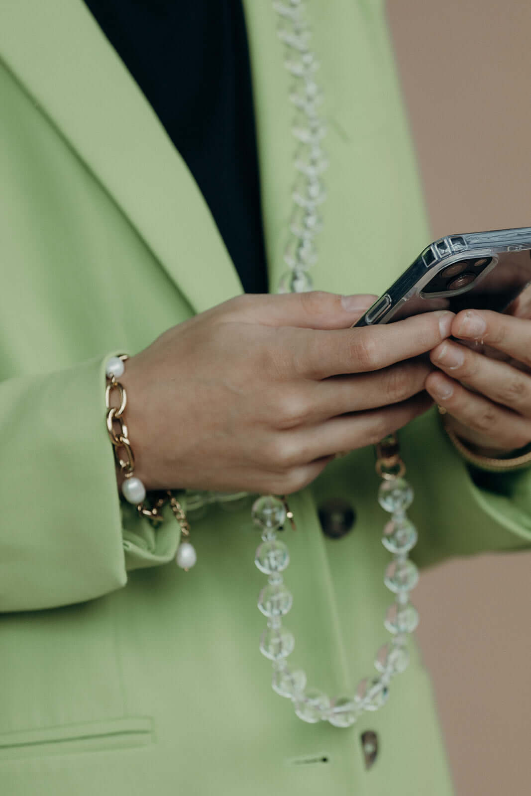 Close-up of hands holding a phone, wearing a gold chain bracelet with pearl charms and a clear glass bead phone strap, over a green blazer.