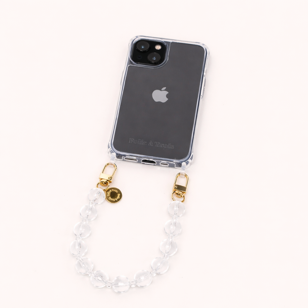 A clear phone case with a clear glass bead phone strap and a gold Folie à Trois charm, shown on a cream background.