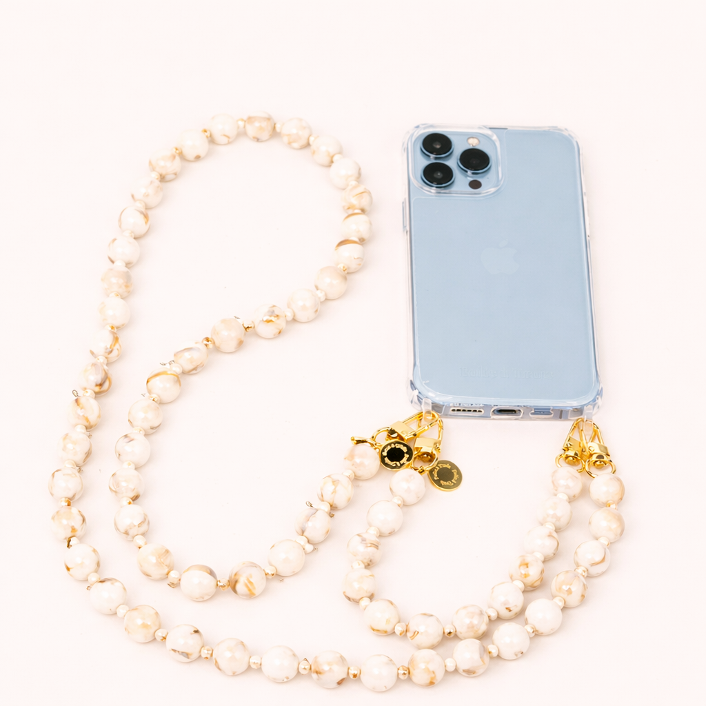 A clear phone case with a cream and gold marbled bead phone strap and gold charms, shown on a cream background.