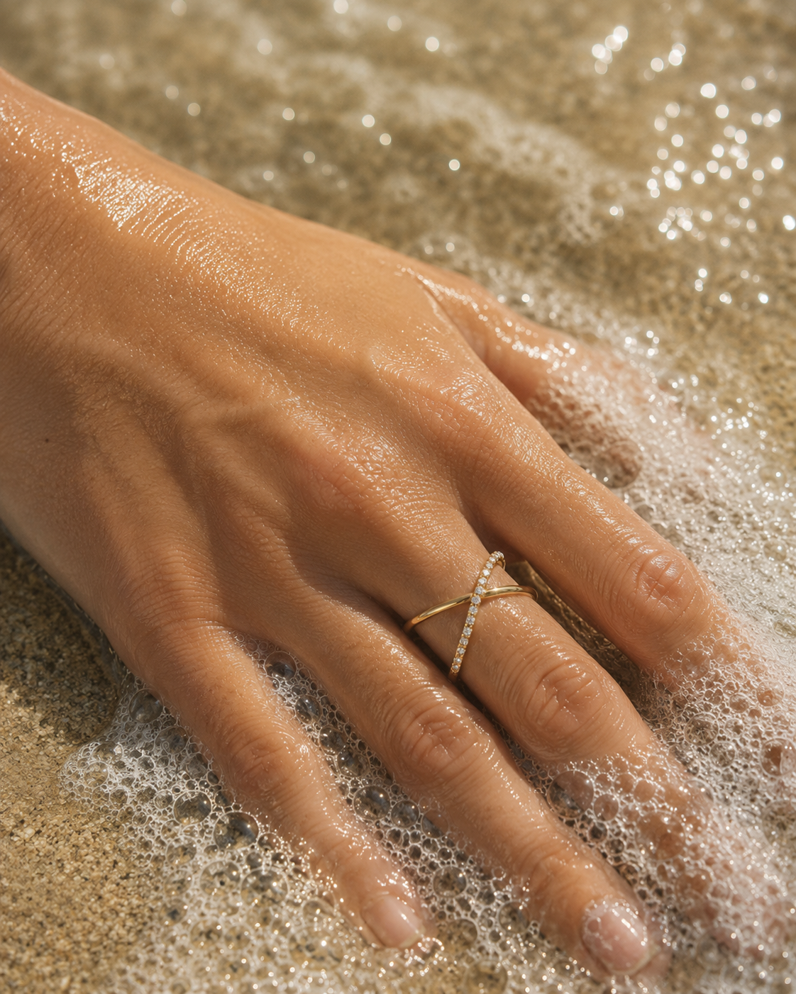 Model wears the gold Kalimera RING on a wet hand at the water's edge, sea foam and sand visible around her fingers.