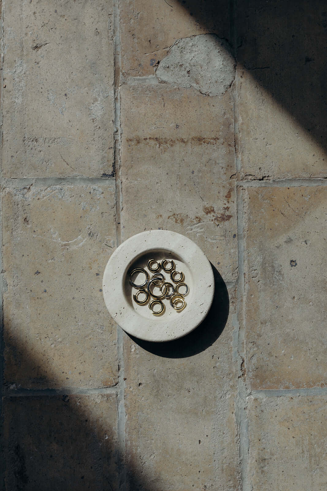 Keep The Base Earrings sit piled in a small stone dish on sunlit tile, their gold hoops catching the light.
