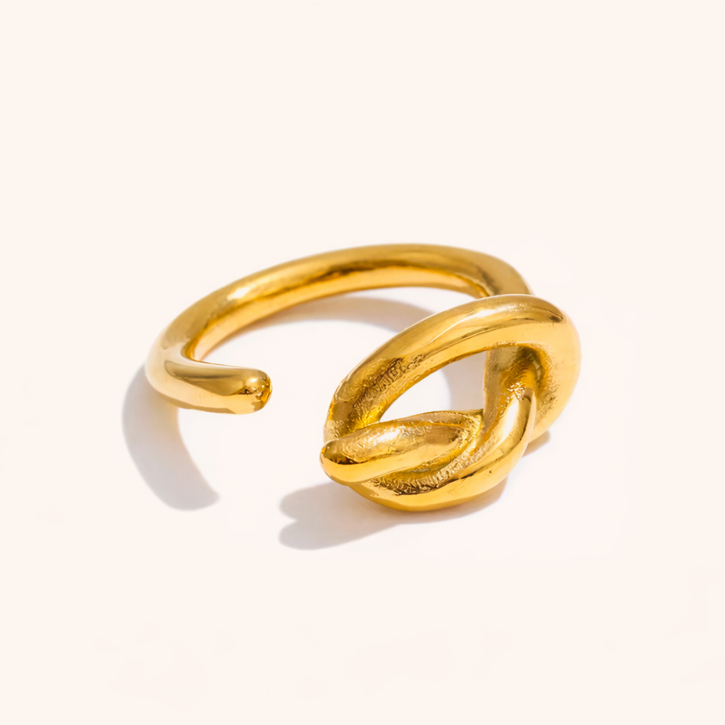 Open gold ring with a sculpted knot detail at the front, shown on a cream backdrop — Folie à Trois Knotted RING.