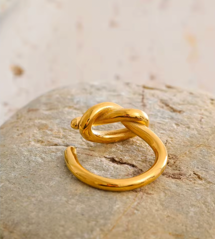 Close-up of the open gold Knotted RING and its sculpted knot resting on a smooth pale stone.