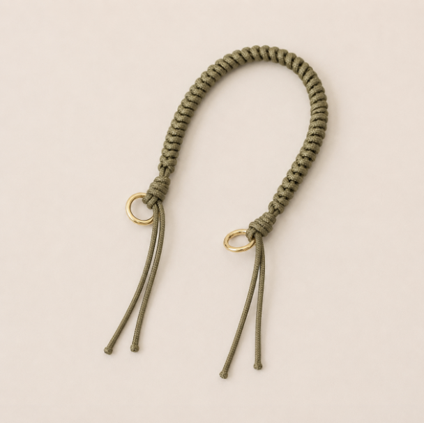 Folie à Trois Layover short wholesale phone strap in khaki green braided cord with gold rings, shown as a knotted loop with fringe ends on a cream background.