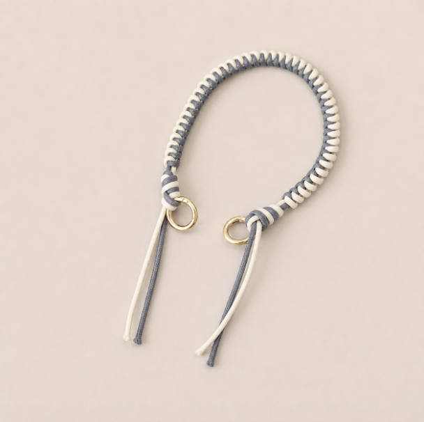 Folie à Trois Layover short wholesale phone strap in gray and white braided cord with gold rings, shown as a knotted loop with fringe ends on a cream background.