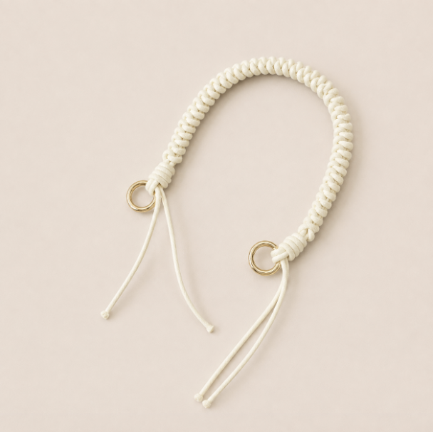 Folie à Trois Layover short wholesale phone strap in white braided cord with gold rings, shown as a knotted loop with fringe ends on a cream background.