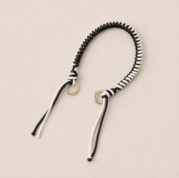 Folie à Trois Layover short wholesale phone strap in black and white braided cord with gold rings, shown as a knotted loop with fringe ends on a cream background.