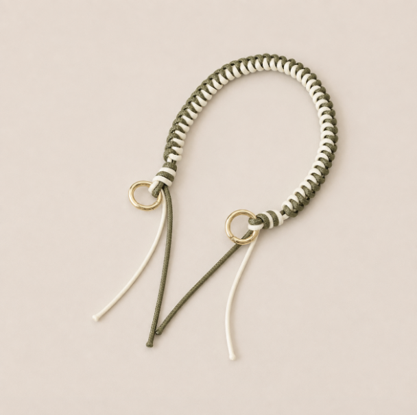 Folie à Trois Layover short wholesale phone strap in green and white braided cord with gold rings, shown as a knotted loop with fringe ends on a cream background.